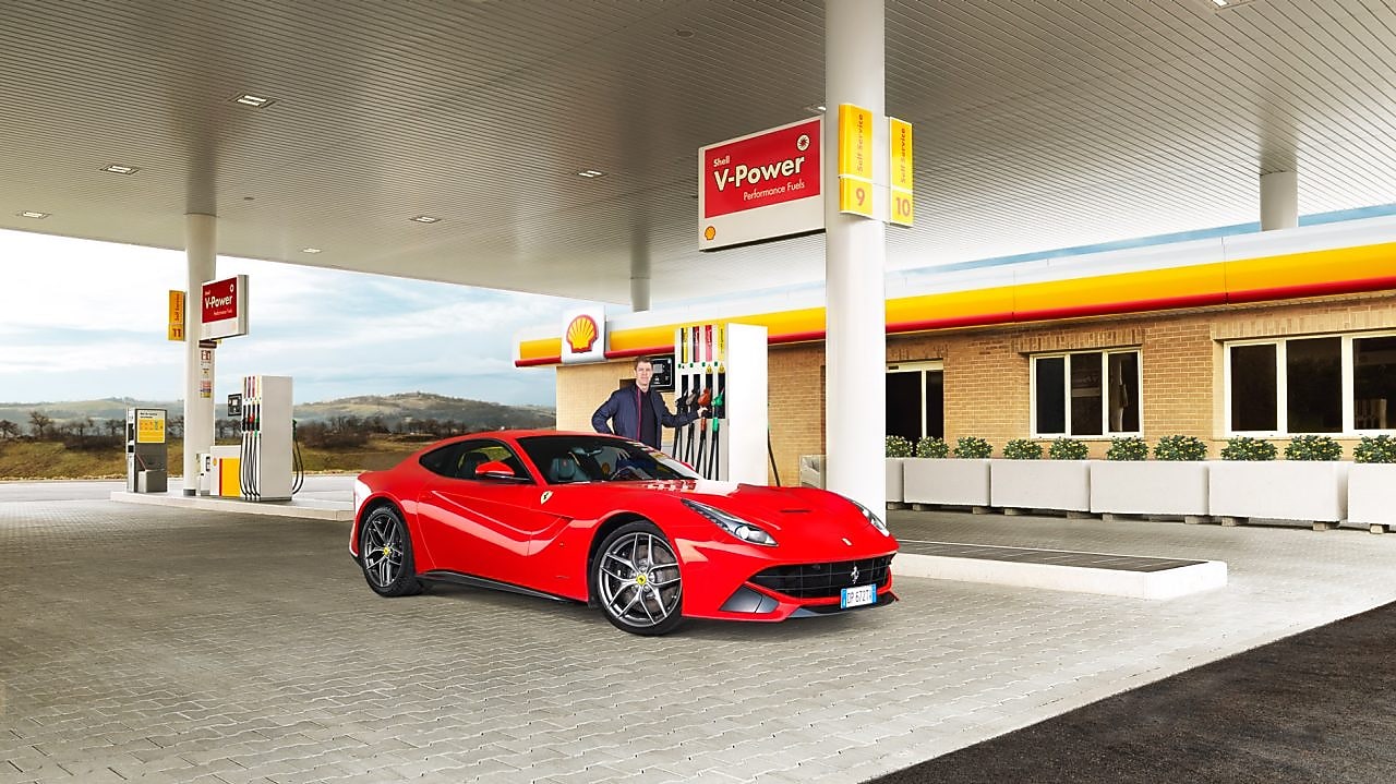 Une Ferrari rouge arrêtée à la pompe d'une station Shell avec un homme qui s'apprête à faire le plein